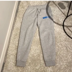 Gråa mjukisbyxor från hoodrich - Gråa mjukisbyxor från hoodrich med broderad blå logga på ena benet. Byxorna har resår och snörning i midjan samt muddar vid bensluten. Tillverkade i mjukt bomullsmaterial för maximal komfort. Herr modell i storlek s men funkar även för tjej då jag användt dem som de
