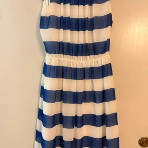 Blåvit randig ärmlös klänning - Snygg ärmlös klänning med breda blå och vita ränder. Modellen har resår i midjan för en skön passform och är tillverkad i ett lätt, luftigt material. Perfekt för dig som gillar en fräsch och somrig stil med lite marint vibe.
