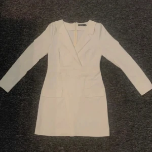 Vit kort klänning från Boohoo - Stilren vit kort klänning från Boohoo med lång ärm och markerad midja. Klänningen har kavajkrage, två fickor framtill och dragkedja i ryggen. Perfekt för dig som gillar en klassisk och elegant look. Aldrig använd!
