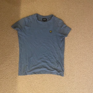 Blå t-shirt från Lyle & Scott Junior - Blå t-shirt från Lyle & Scott Junior med klassisk rund hals och korta ärmar. T-shirten har en gul broderad logga på bröstet och är tillverkad i mjuk bomull. Perfekt för en avslappnad stil.