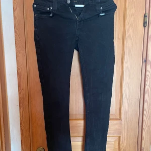 Svarta jeans från Nudie Jeans - Svarta jeans från Nudie Jeans med klassisk femficksdesign och diskret broderi på bakfickorna. Jeansen har smal passform och raka ben, tillverkade i mjukt bomullstyg med stretch för extra komfort. Perfekta för dig som gillar stilrena och tidlösa jeans.