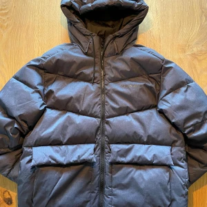 Mörkgrå pufferjacka Jack & Jones - Pris kan diskuteras! Snygg mörkblå pufferjacka från Jack & Jones Originals med huva och stora fickor framtill. Jackan har dragkedja och är perfekt för kalla dagar. Den har en clean look och är riktigt skön att bära med sin vadderade design.