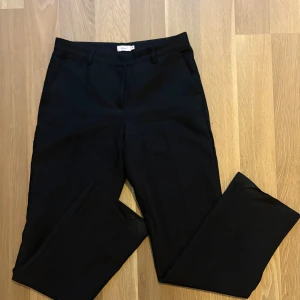 Svarta kostymbyxor från Nelly - Klassiska svarta kostymbyxor från Nelly i storlek 36. Byxorna har raka ben, hällor för bälte och diskreta fickor både fram och bak. Perfekta för en clean och stilren look. Materialet känns mjukt och följsamt.