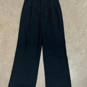 Svarta vida kostymbyxor - Snygga svarta kostymbyxor med hög midja och vida ben. Klassisk design med hällor för bälte och en diskret bakficka. Perfekta för en clean och stilren look. Materialet känns slätt och har en lätt glans.