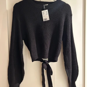 Svart stickad tröja med knyt från H&M - Svart stickad tröja från H&M med croppad passform och snygg knytning bak. Tröjan har rund halsringning och långa ärmar med ribbade muddar. Perfekt för lager-på-lager och passar till både jeans och kjol.