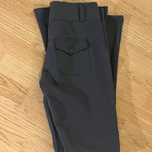 Gråa bootcut-byxor från Gina Tricot - Gråa bootcut-byxor från Gina Tricot i stretchigt material. Byxorna har hög midja, bälteshällor och en ficka med knapp bak. Perfekta för dig som gillar en stilren och trendig look.