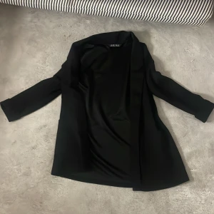 Svart kavaj från Amisu, storlek 36 - Svart kavaj från Amisu i stilren design med lång ärm och klassisk krage. Materialet är mjukt och har en lätt glansig finish, vilket ger en snygg look. Perfekt för dig som gillar minimalistisk stil och vill ha en enkel svart kavaj i garderoben.