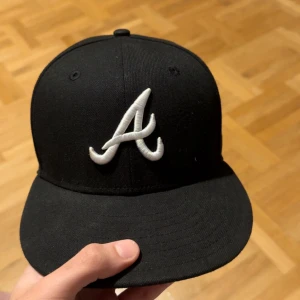 Svart New Era 59FIFTY keps Atlanta - Snygg svart New Era 59FIFTY keps med Atlanta Braves A-logga i vitt framtill och New Era-logga på sidan. Klassisk flat peak och stilren look, perfekt för dig som gillar streetwear. Tillverkad i bomull och polyester för skön passform.