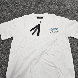 Vit AMIRI t-shirt med blommor - Vit t-shirt från AMIRI med blå logga och blommigt tryck både fram och bak. Klassisk passform med rund hals och korta ärmar. Tillverkad i mjuk bomull, perfekt för en clean och stilren look.
