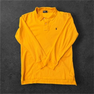 Gul långärmad pikétröja från Ralph Lauren - Snygg långärmad pikétröja från Polo Ralph Lauren i en stark gul färg. Klassisk krage med två knappar och broderad logga på bröstet. Tillverkad i mjuk bomull som känns skön mot huden. Perfekt för dig som vill sticka ut lite extra.