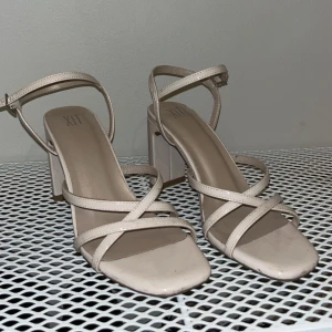 Beige klackskor från XIT - Snygga beige sandaler från XIT med höga blockklackar och smala remmar som korsar över foten. Skorna har öppen tå och justerbart spänne runt ankeln. Perfekta för dig som gillar stilrena och trendiga skor med lite extra höjd. 