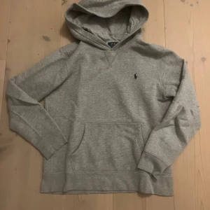 Grå hoodie från Polo Ralph Lauren - Snygg grå hoodie från Polo Ralph Lauren med klassisk broderad logga på bröstet. Tröjan har huva, känguruficka och långa ärmar. Perfekt för chill dagar och enkel att matcha med jeans eller joggers.