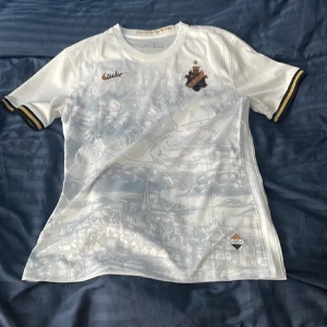 AIK vit kortärmad fotbollströja Nike - AIK:s vita kortärmade fotbollströja från Nike med subtilt stadsmotiv i ljusgrått över hela framsidan. Tröjan har svarta och gula detaljer på ärmsluten, AIK:s klubbmärke på bröstet och Nike-logga i guld. Materialet är lätt och andas, perfekt för match eller träning.