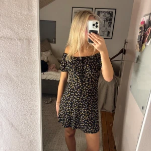 Svart blommig offshoulder klänning - Supersöt svart kortklänning med små blommor i vitt och gult. Offshoulder-modell med korta ärmar och lätt rynk vid halsen.