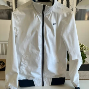 Vit vindjacka från Etirel med dragkedja - Fräsch vit vindjacka från Etirel med svart detaljerad dragkedja och logga på bröstet. Jackan har ribbade muddar och är tillverkad i en mix av polyester och bomull. Perfekt för dig som gillar stilren och sportig look.