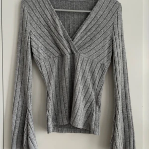 Grå ribbad omlott-tröja med v-ringning - Grå ribbad tröja med omlott-design och djup v-ringning. Tröjan har långa ärmar med lätt utsvängd form. Bara användts 1 gång. Skriv om du har frågor💗