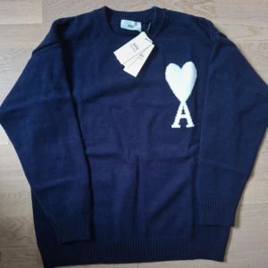 Marinblå stickad tröja från Ami - Snygg marinblå stickad tröja från Ami Alexandre Mattiussi med vit hjärt- och A-logga på bröstet. Rund hals och ribbade muddar vid ärmslut och nederkant. Perfekt för dig som gillar stilrena detaljer och vill ha något som sticker ut.