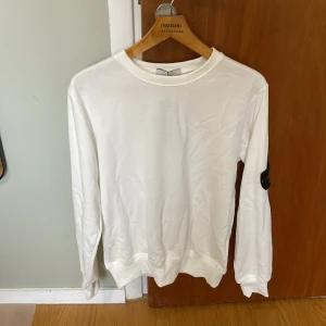 Vit sweatshirt från Stone Island - Vit långärmad sweatshirt från Stone Island med klassisk rund hals och ikonisk logotyp-patch på vänster ärm. Tröjan är i mjuk bomull och har ribbade muddar vid ärmslut och nederkant. Perfekt för en clean och stilren look.