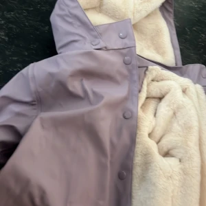 Ljuslila regnjacka med teddyfoder Stl 146 - Snygg ljuslila regnjacka med huva och mjukt, krämfärgat teddyfoder på insidan. Jackan har tryckknappar framtill och två fickor. Perfekt för kalla och regniga dagar när du vill vara både varm och stilren. Aldrig använd , Stl 146