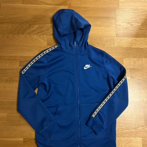 Blå Nike hoodie med huva och dragkedja - Snygg blå hoodie från Nike med huva och hel dragkedja framtill. Tröjan har vita Nike-loggor längs axlarna och ett tryckt Nike-märke på bröstet. Tillverkad i mjuk polyester med praktiska fickor framtill. Perfekt för chill eller träning.
