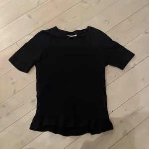 Svart blus med volang från Gina Tricot - Svart blus från Gina Tricot med korta ärmar och rund hals. Blusen har en snygg volangkant nedtill och är tillverkad i ett lätt, syntetiskt material. Perfekt för dig som gillar stilrena plagg med en liten twist. Supersnygg att matcha med jeans eller kjol.