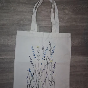 Vit tygkasse med blommigt tryck - En stilren vit shoppingväska i ett tjockt material med ett blommigt tryck i blått, lila och gult på ena sidan. Perfekt för att bära böcker, laptop eller shopping. Enkel och trendig design som passar till många outfits. Mått ca 34x39cm