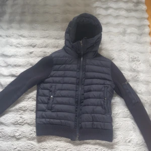 Svart Moncler pufferjacka med stickade ärmar - Snygg svart pufferjacka från Moncler med stickade ärmar och huva. Jackan har dragkedja framtill, två fickor med dragkedja och en extra ficka på ärmen. Kombinerar quiltad look med stickade detaljer för en modern vibe. 