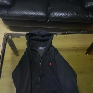 Svart hoodie från Polo Ralph Lauren - Svart hoodie från Polo Ralph Lauren med röd broderad logga på bröstet. Tröjan har huva med snörning och dragkedja framtill. Tillverkad i mjuk bomull, perfekt för chill dagar. Klassisk och stilren design som funkar till jeans eller joggers.
