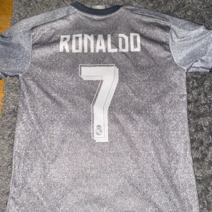Real Madrid Ronaldo 7 bortatröja - Säljer en grå Real Madrid bortatröja från Adidas med Ronaldo 7 på ryggen. Tröjan har korta ärmar, gula ränder på axlarna och klubbmärke samt Fly Emirates-tryck på bröstet. Tillverkad i lätt och ventilerande material, perfekt för fotboll eller samling. Och är runt 150 i storlek 