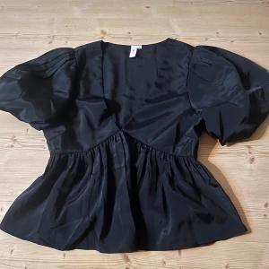 Svart blus med puffärmar - Säljer denna super snygga blusen med puffiga ärmar, v-ringning och volang längst ner. Jätte fint glansigt material. 🩷🩷