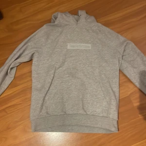 Grå hoodie från Peak Performance - Snygg grå hoodie från Peak Performance med broderad logga på bröstet. Klassisk modell med huva och känguruficka framtill. Mjuk och skön i bomullsblandning, perfekt för chill eller skola.