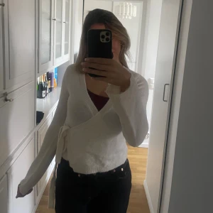 Vit omlottkofta med knytband - Mysig vit omlottkofta med knytband i sidan och v-ringad hals. Koftan har lång ärm och är stickad i ett mjukt material. Perfekt att slänga på sig över ett linne eller t-shirt för en chill look.