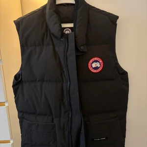Svart dunväst från Canada Goose | KVITTO FINNS! - Svart dunväst från Canada Goose med klassisk logotyp på bröstet. Västen har hög krage, två stora fickor framtill och stängs med dragkedja och tryckknappar. Perfekt för kalla dagar och har en stilren, quiltad design. KVITTO FINNS!