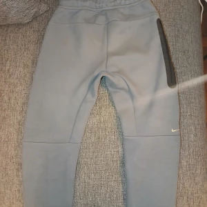 Grå Nike mjukisbyxor storlek L - Snygga grå mjukisbyxor från Nike med gul logga på vänster höft och svart dragkedja på ena benet. Byxorna har resår och snörning i midjan samt praktiska fickor. Perfekta för chill eller träning, tillverkade i mjukt bomullsmaterial.