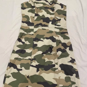 Camouflage klänning med dragkedja - Säljer en klänning med klassiskt camouflage-mönster i grönt, beige, svart och brunt. Klänningen har figurnära passform och dragkedja i ryggen. Perfekt för dig som vill sticka ut med en edgy stil.