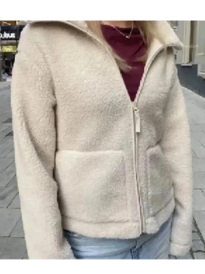 Beige teddyjacka med dragkedja - Mysig beige teddyjacka med hög krage och två stora fickor framtill. Jackan har en rak passform och stängs med dragkedja. Perfekt för dig som gillar chill och comfy stil, funkar fett bra till jeans eller mjukis. Materialet är fleece som ger en fluffig och varm känsla.