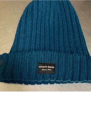 Grön/blå  stickad mössa från North Bend - Snygg blå/grön  ribbstickad mössa från North Bend med uppvikt kant och svart logolapp framtill. Tillverkad i 100% akryl, perfekt för att hålla värmen under kyliga dagar. Enkel och stilren design som passar till de flesta jackor. Fri frakt 