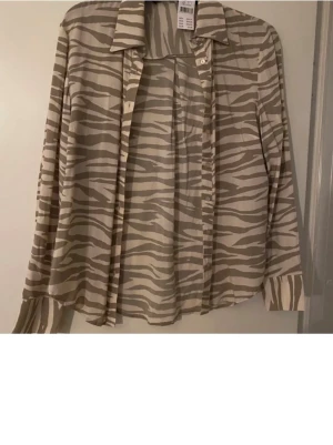 Beige zebramönstrad skjorta strl S  - Säljer en beige skjorta från KappAhl med zebramönster i ljusbruna toner. Skjortan har klassisk krage, knappar framtill och långa ärmar. Materialet är mjukt och känns som bomull, perfekt för dig som gillar djurmönster och vill sticka ut lite extra. Ny med prislapp 