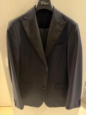 Marinblå smoking rån Oscar Jacobson - Snygg blå smoking från Oscar Jacobson med breda slag i satin och två knappar framtill. Klassisk design med fickor och elegant passform, perfekt för dig som vill ha en stilren look. Materialet är slätt och har en lyxig känsla.