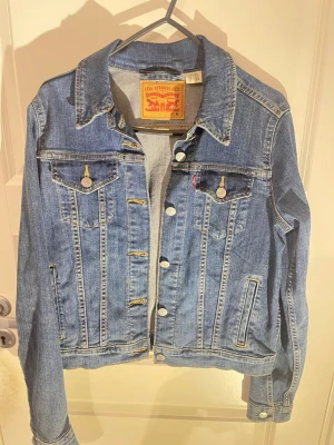 Levi's klassisk jeansjacka blå M - Säljer en klassisk blå jeansjacka från Levi's i storlek M. Jackan har silverfärgade knappar, två bröstfickor med lock och knapp samt två sidofickor. Snygga sömmar och tidlös passform, perfekt till alla dina outfits.