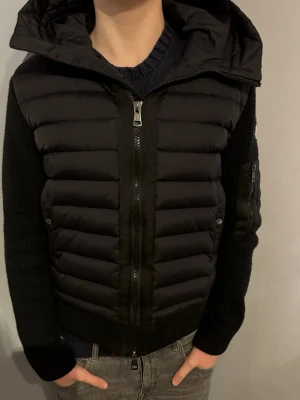 Moncler cardigan - Säljer en svart pufferjacka från Moncler med stickade ärmar och huva. Jackan har dragkedja framtill, fickor med dragkedja på ärmen och Moncler-logga på armen. Kroppen är quiltad och vadderad medan ärmarna är ribbstickade för en schysst kontrast.