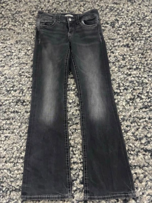 Bootcut jeans från Gina Tricot, svart - Svarta bootcut jeans från Gina Tricot i storlek 158. Snyggt tvättade med ljusare partier och kontrastsömmar. Klassisk femficksmodell med knappar och dragkedja framtill samt fickor med lock och knapp bak. Perfekt för dig som gillar en cool och avslappnad stil.