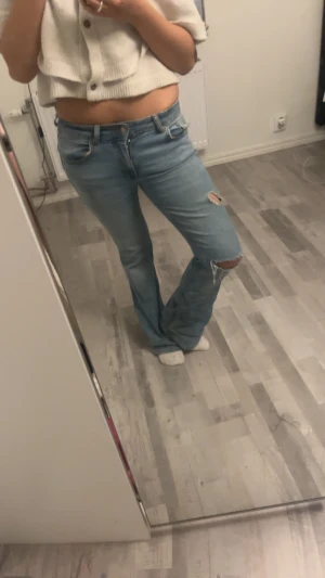 Bootcut ljusblå jeans med slitningar - Säljer ett par ljusblå bootcut jeans med snygga slitningar och hål på ena benet. Jeansen har klassisk femficksdesign, låg midja och är tillverkade i mjukt denimtyg. Perfekt för dig som gillar en avslappnad och trendig look.