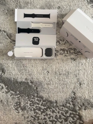 Apple Watch  - Apple Watch med svart och vitt armband, USB-C laddkabel och originalförpackning. Klockan ser ut att vara i mycket gott skick utan synliga repor eller skador. Perfekt för träning, hälsa och smarta funktioner. Komplett med tillbehör.