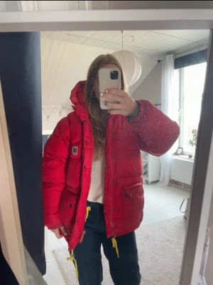 Röd pufferjacka från Fjällräven - Säljer en röd pufferjacka från Fjällräven med stor huva och broderad logga på bröstet. Jackan har flera fickor med knappar, dragkedja framtill och quiltad design. Perfekt för kalla dagar och har en riktigt schysst oversized passform. 