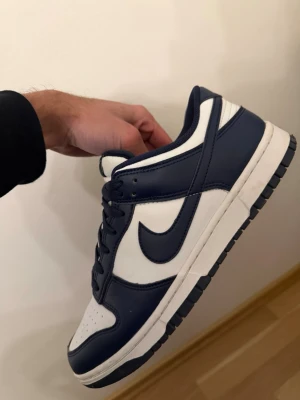 Nike Dunk Low blå och vita sneakers - Säljer ett par Nike Dunk Low sneakers i marinblått och vitt. Skorna har klassisk låg siluett, snörning och rund tå. Ovandelen är i skinn med kontrasterande färgblock och den ikoniska swoosh-loggan på sidan. Perfekta för dig som gillar streetwear och vill ha en clean look.