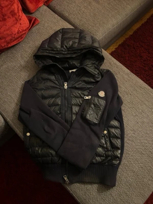 Moncler cardigan med huva - Säljer en marin blå cardigan str XL men skulle säga att det är M från Moncler med quiltade partier och stickade ärmar. Jackan har en stor huva, dragkedja framtill och en praktisk ficka med dragkedja på ärmen. Klassisk Moncler-logga på armen och ribbade muddar för extra komfort.  Priset är diskuterbart, jag är bara ett meddelande bort. Svarar alltid så fort jag kan.