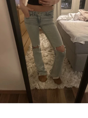 Ljusblå bootcut jeans med hål - Säljer ett par ljusblå jeans med bootcut-snitt och slitna hål på båda knäna. Jeansen har klassisk femficksdesign och broderad detalj på bakfickan. Snyggt lågmidjade och tillverkade i jeansmaterial. Perfekta för dig som gillar en chill och trendig look.