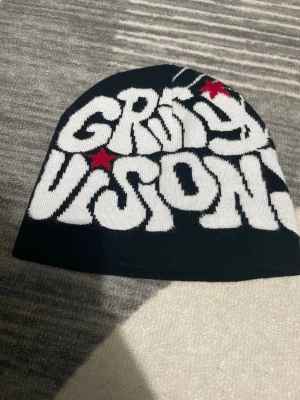 Svart stickad mössa med tryck - Cool svart stickad mössa från Grimy Vision med grafiskt vitt tryck och röda stjärnor på ena sidan, samt ett stort spindelnätsmönster i vitt på andra sidan. Perfekt för dig som gillar streetwear och vill sticka ut med din stil.