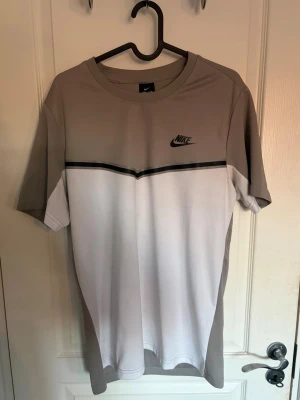 Nike t-shirt i beige och vit - Snygg t-shirt från Nike i beige och vit med svart logga på bröstet. T-shirten har en cool färgblock-design med en svart linje över bröstet och klassisk rund hals. Perfekt för dig som gillar sportig och stilren look.